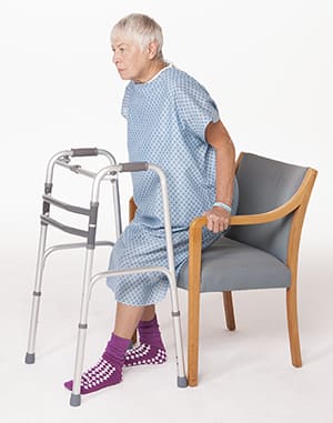 Person in hospital gown with walker in front of them, holding onto arms of chair and safely lowering self down in chair.
