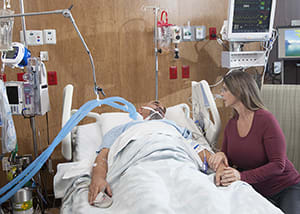 Person sitting next to bed of intubated patient in intensive care unit.